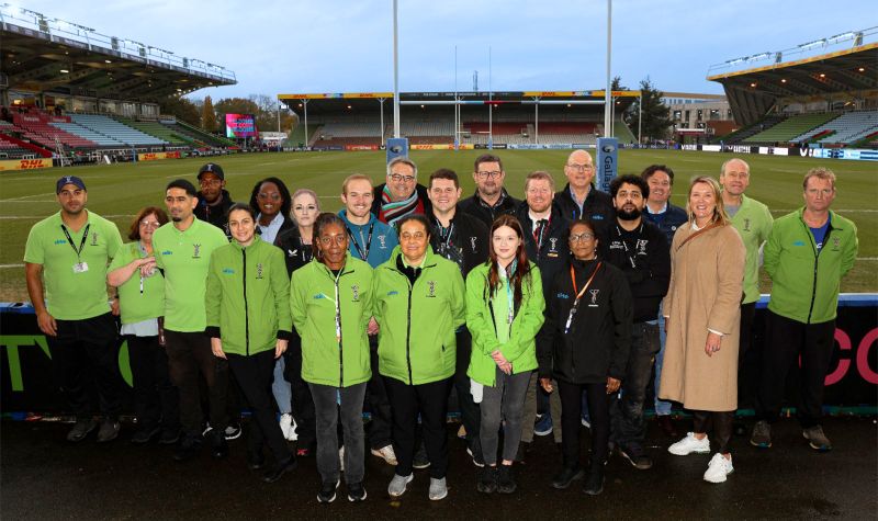 A group of people stand in front of a rugby oval. Half of them are wearing fluorescent green jackets with the Cirka symbol. The other half are wearing black jackets with Cirka branded on them.