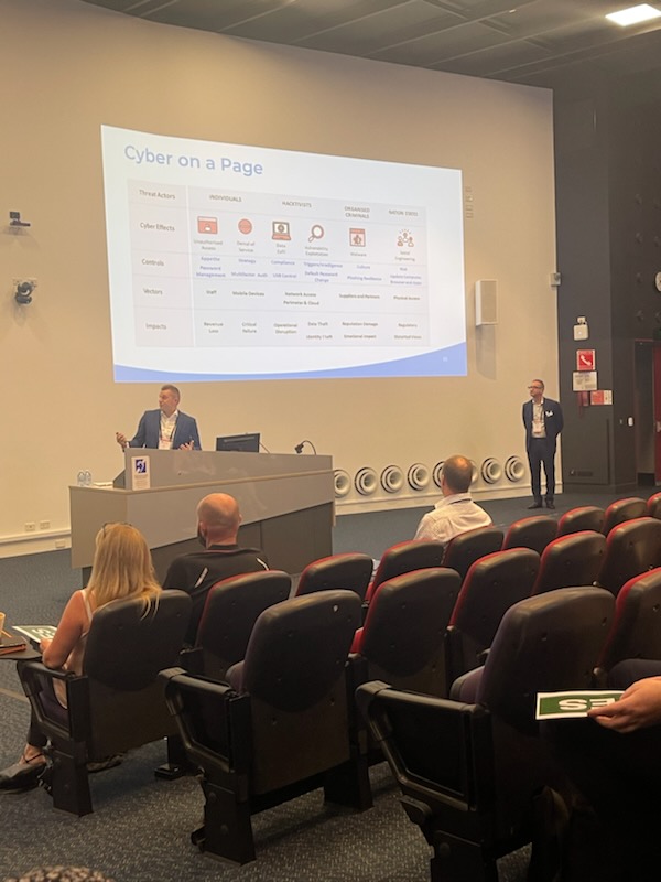 Man stands in front of a presenting stand in a lecture theatre. People in the audience are watching him. A presentation on cyber security is presented behind him.
