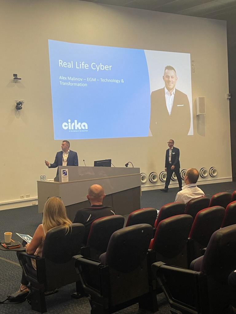 Two men stand in front of a presenting stand in a lecture theatre. People in the audience are watching him. A presentation on cyber security is presented behind him.