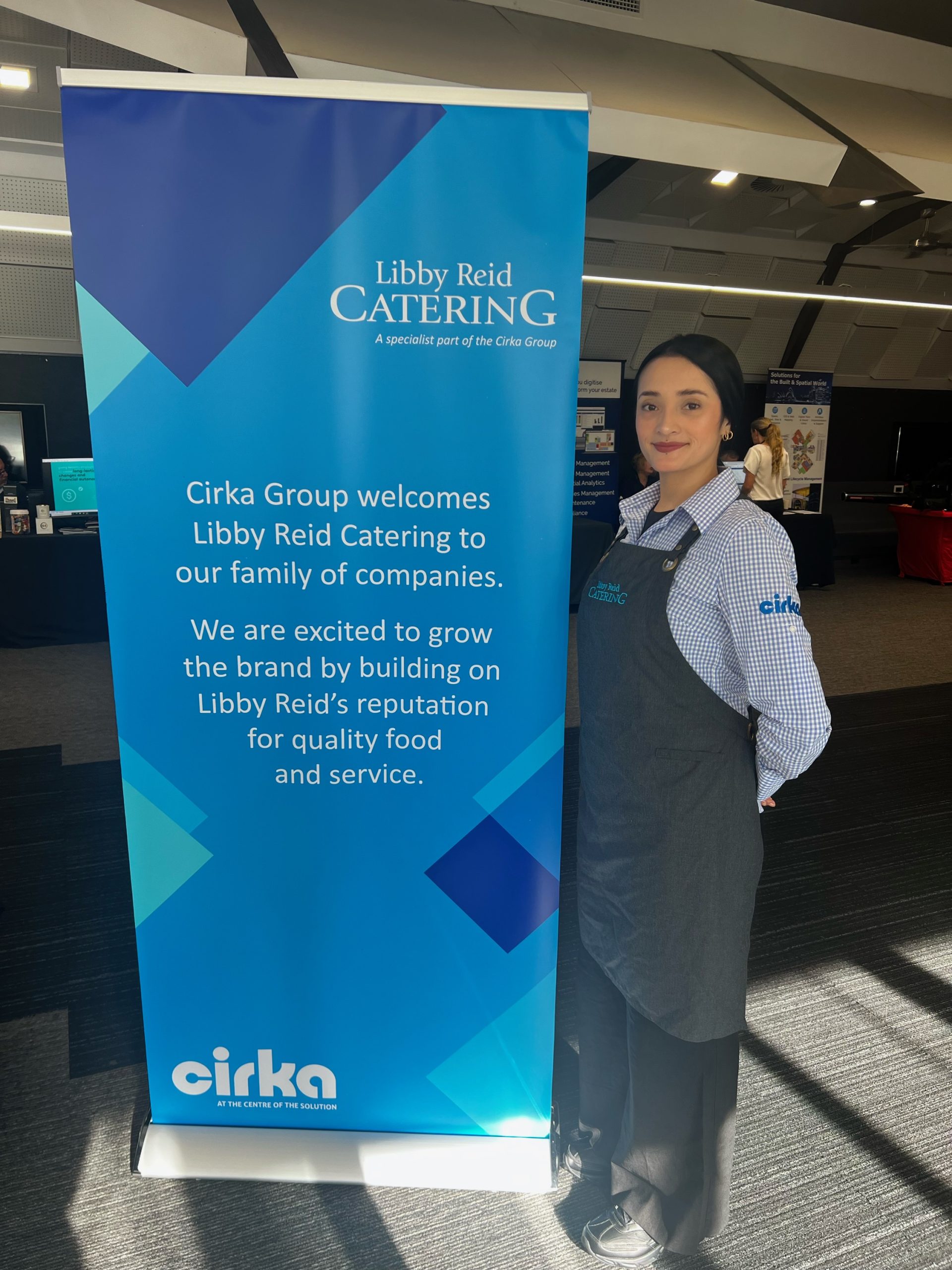 Woman stands in front of a Libby Reid Catering sign that welcomes Libby Reid Catering to Cirka Group. The woman wears an apron with Libby Reid Catering on it and a patterned shirt with Cirka on the arm band