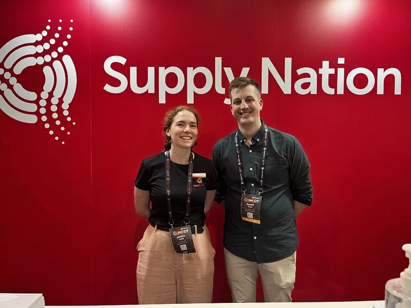 1 female-presenting and 1 male-presenting person stand in front of a large red logo that reads SupplyNation.